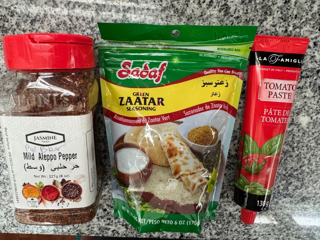 photo showing three packages of food: a dark red ground pepper labelled "mild Aleppo Pepper" with the brand name "Jasmine," a bag  labelled "green zaatar seasoning" with the brand name "Sadaf," and a tube of tomato paste with the brand name "la famiglia."