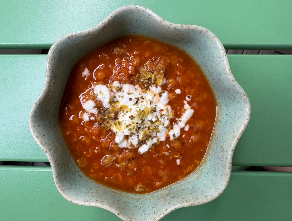 a photo of a bowl of soup. It is tomato red with lentils in it, and has crumbled goat cheese, herbs and lemon zest on top.